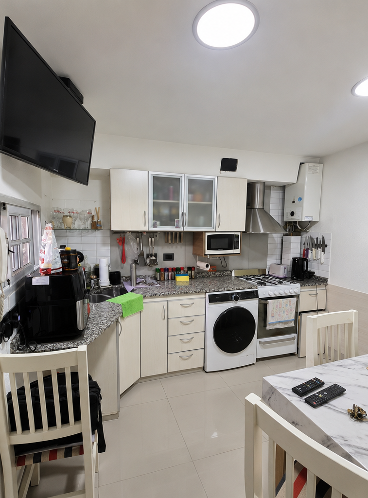 Cocina compacta con armarios blancos, lavadora, estufa, televisor montado en la pared y encimeras desordenadas.