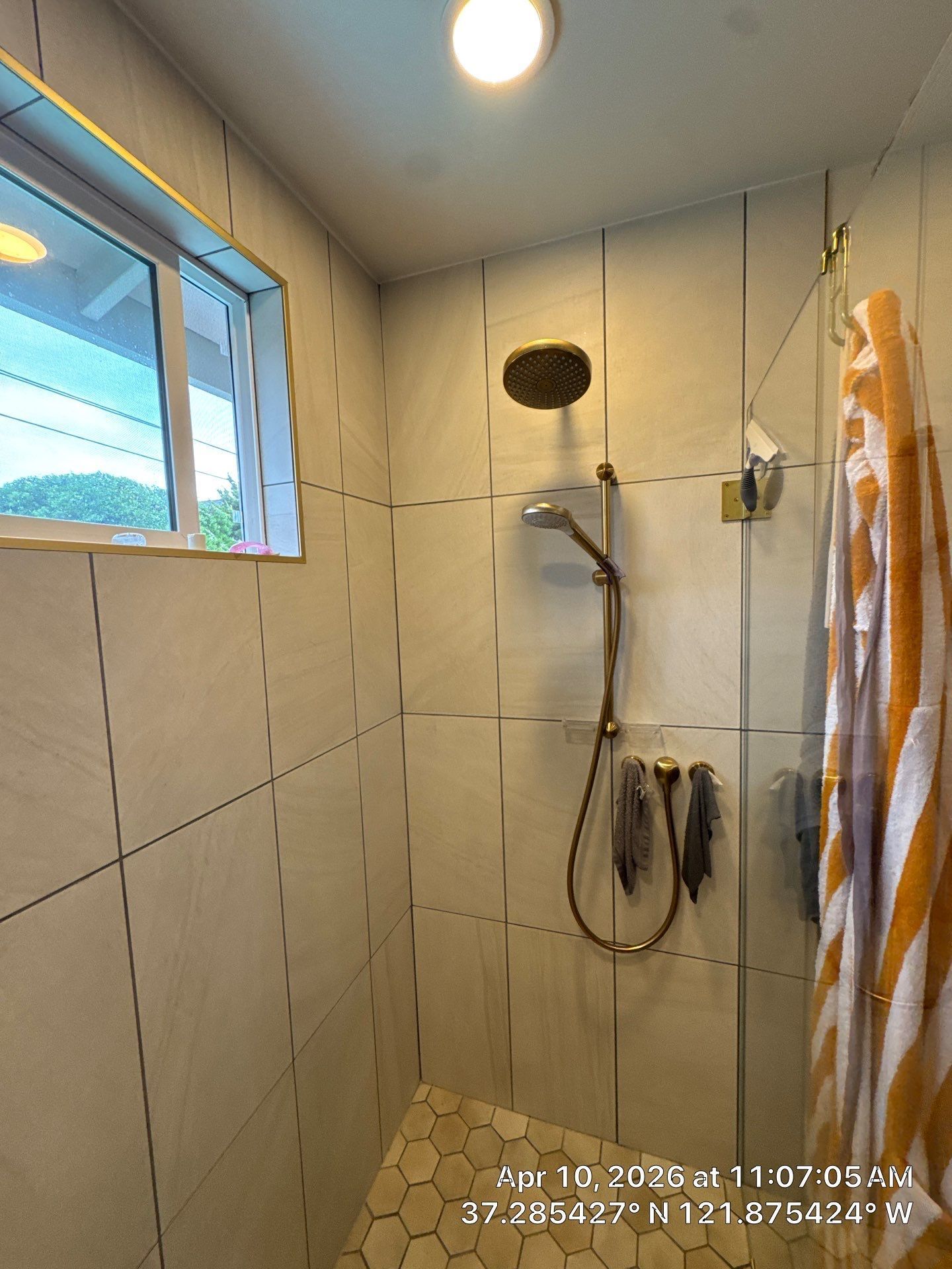 Small tiled shower stall with overhead light, window, handheld showerhead, and striped towel on the right.