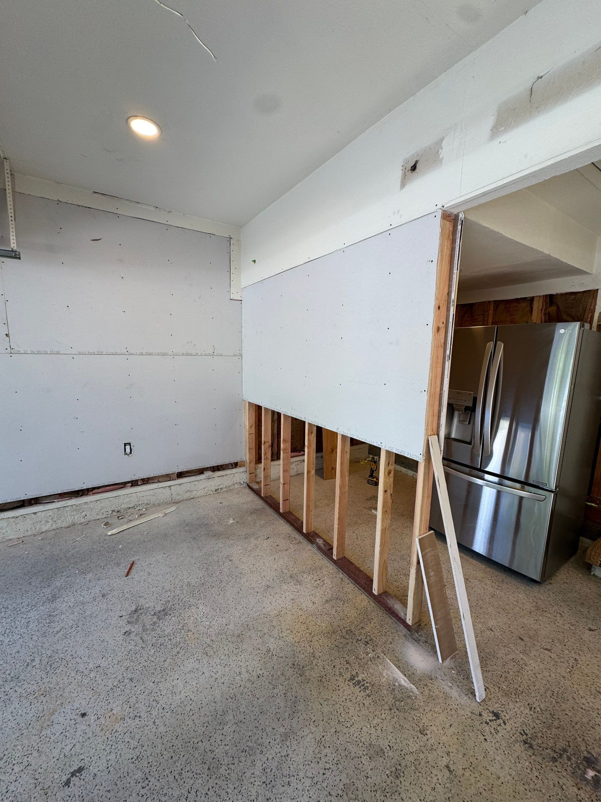 Partially demolished kitchen with exposed studs, white walls, and a stainless steel refrigerator in the background