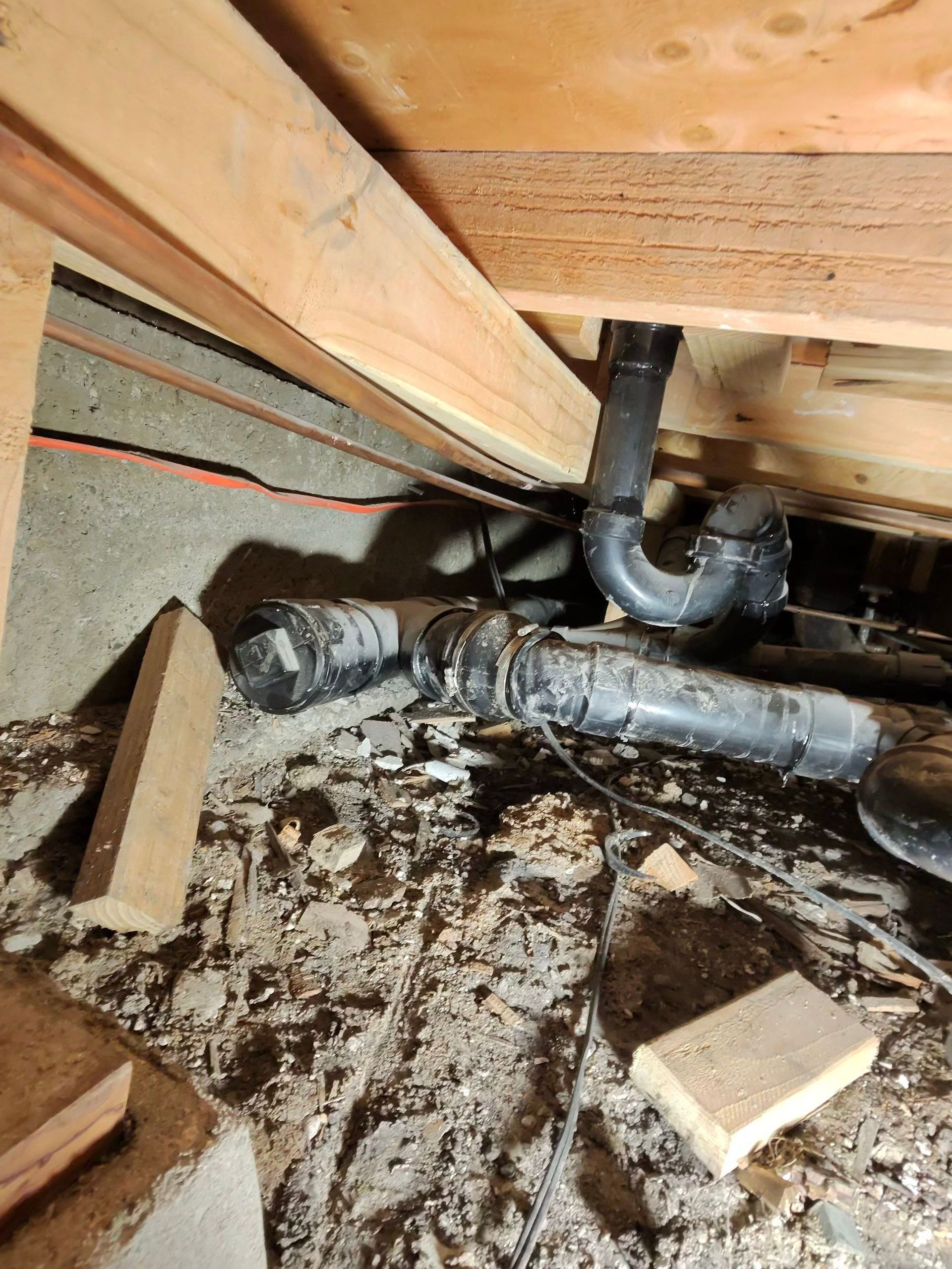 Crawlspace plumbing with black pipes, wood framing, insulation, and scattered debris on the ground.