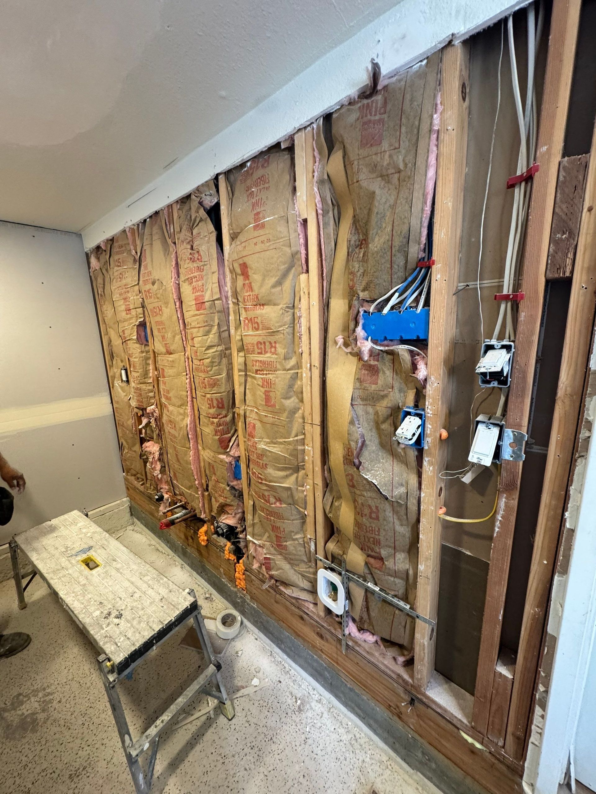 Exposed wall studs and insulation during home renovation, with a ladder in front and tools mounted on the wall.