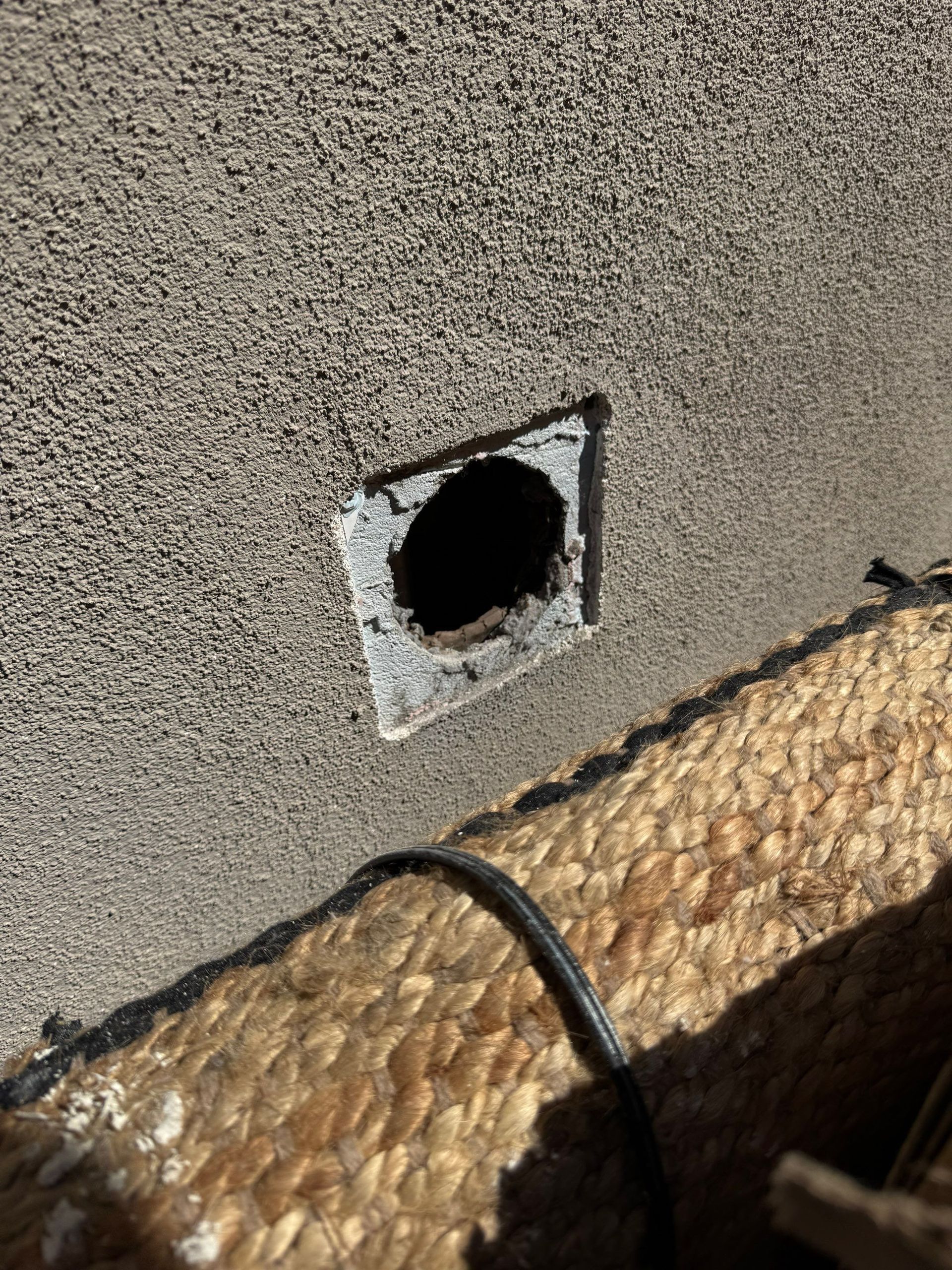 Exterior wall vent opening beside a woven mat or thatched surface, with a black cable hanging down