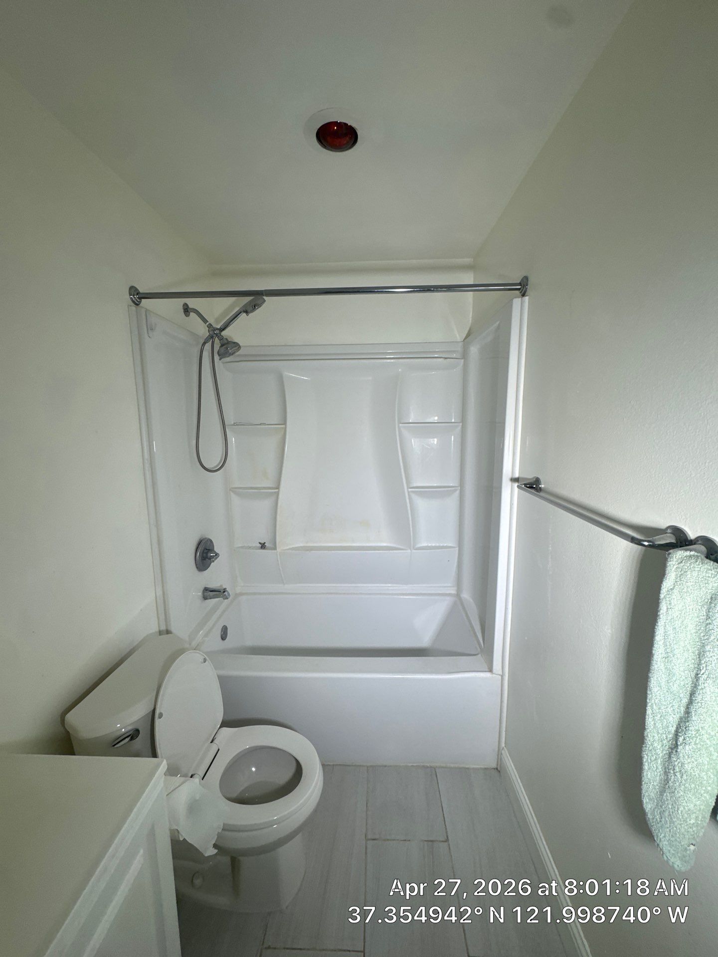 Small bathroom with toilet and shower tub, white walls, towel rack, and a red ceiling smoke detector.