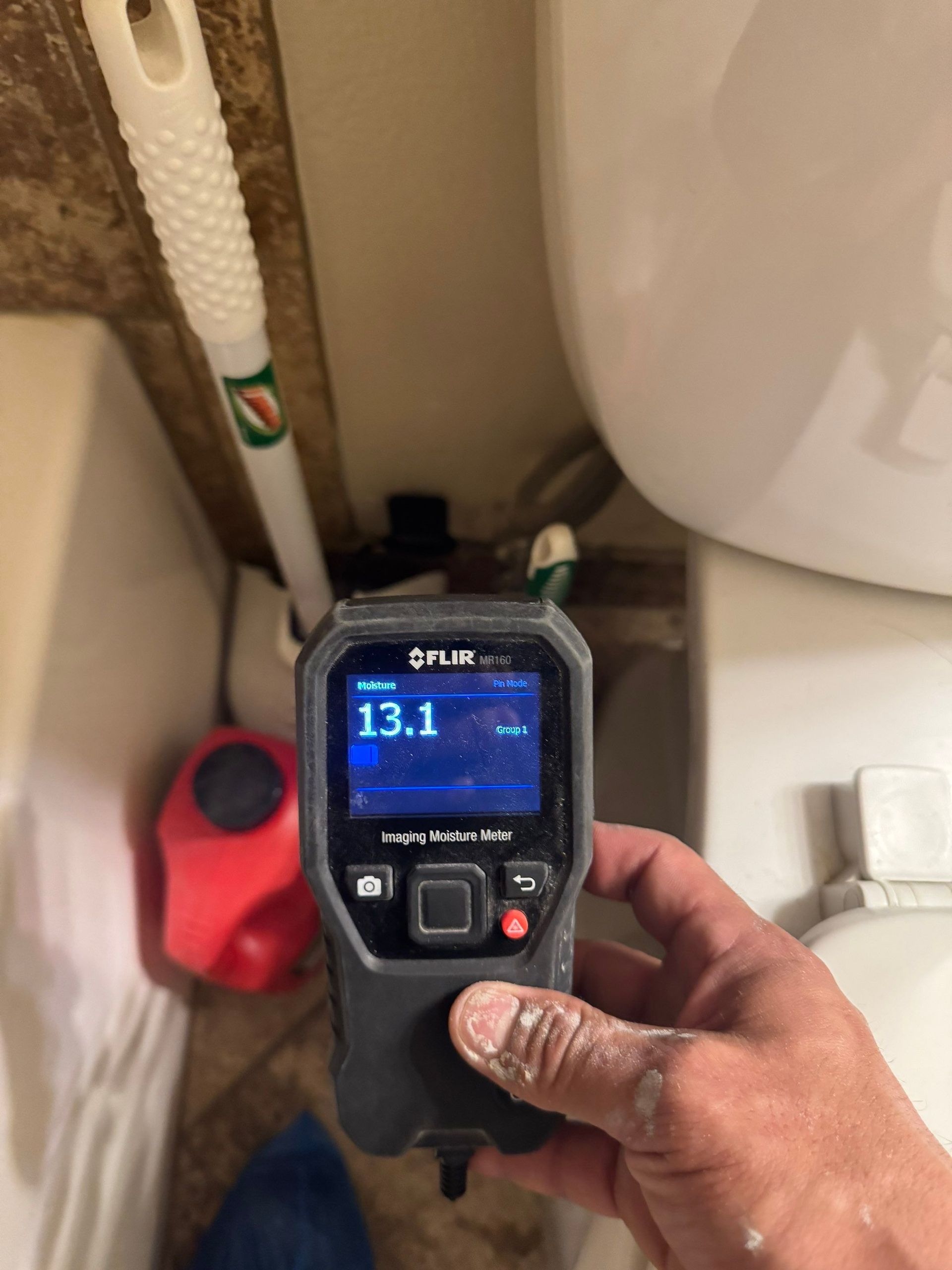 Hand holding a digital meter reading 13.1 near a toilet and plumbing in a bathroom/utility space
