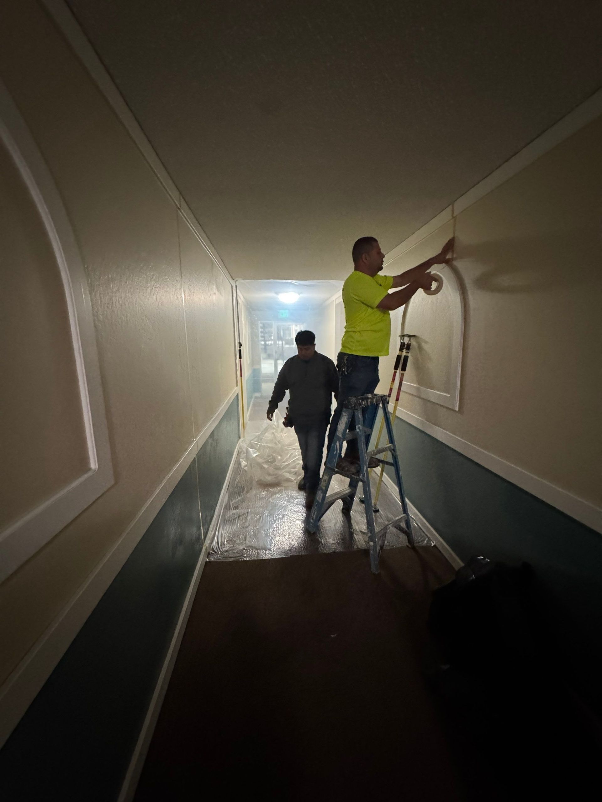 Two people painting a narrow hallway, one on a ladder, with plastic-covered floor and bright light ahead