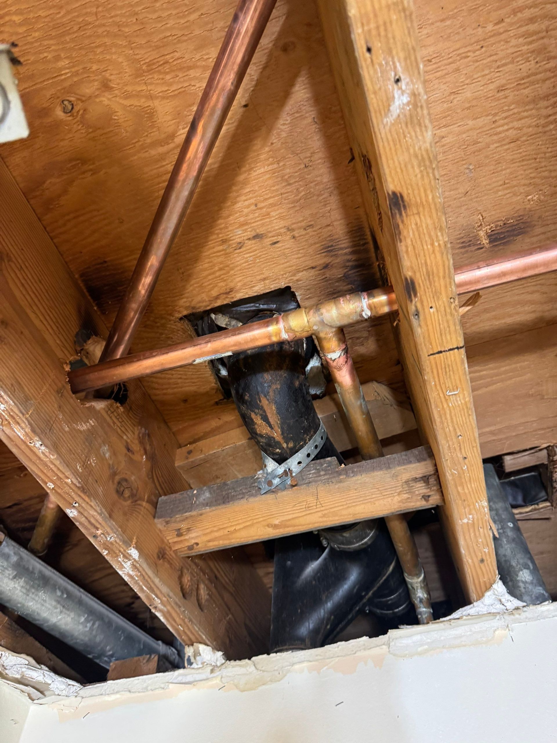 Exposed copper pipes and a black plumbing vent beneath a damaged wooden floor and joists
