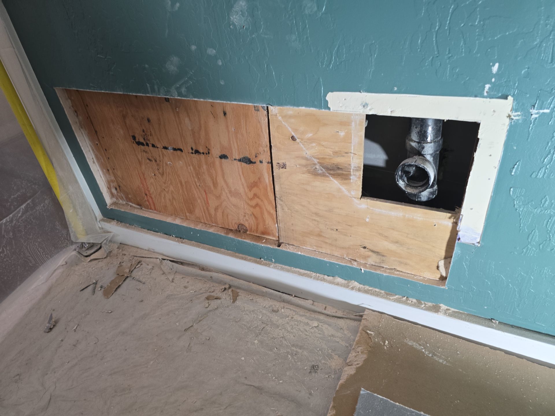 Partially covered wall opening with exposed pipe and plywood patch in a teal room