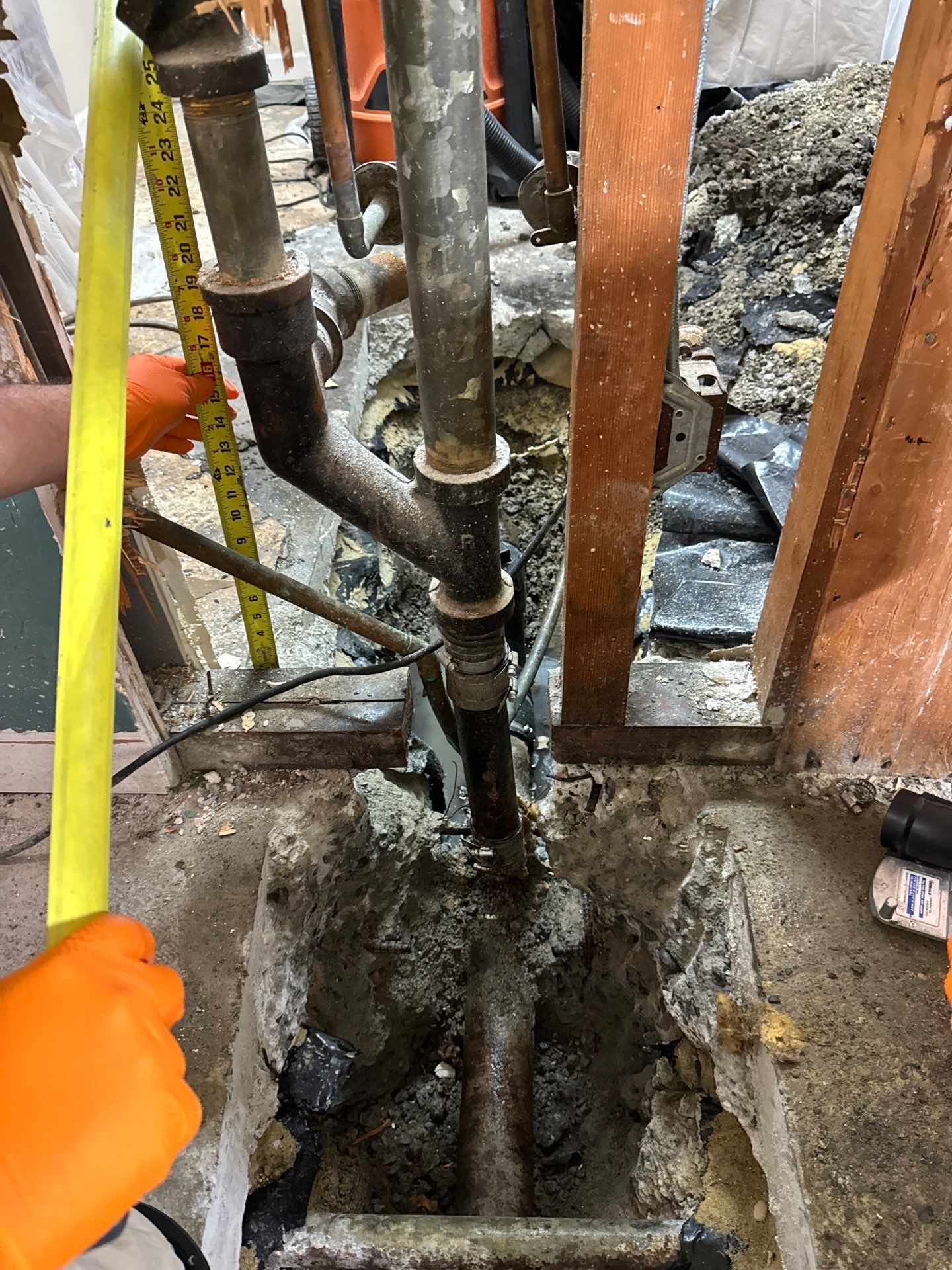 Plumbing pipes and a worker’s orange-gloved hand inspecting a wet, damaged floor area in a crawl space