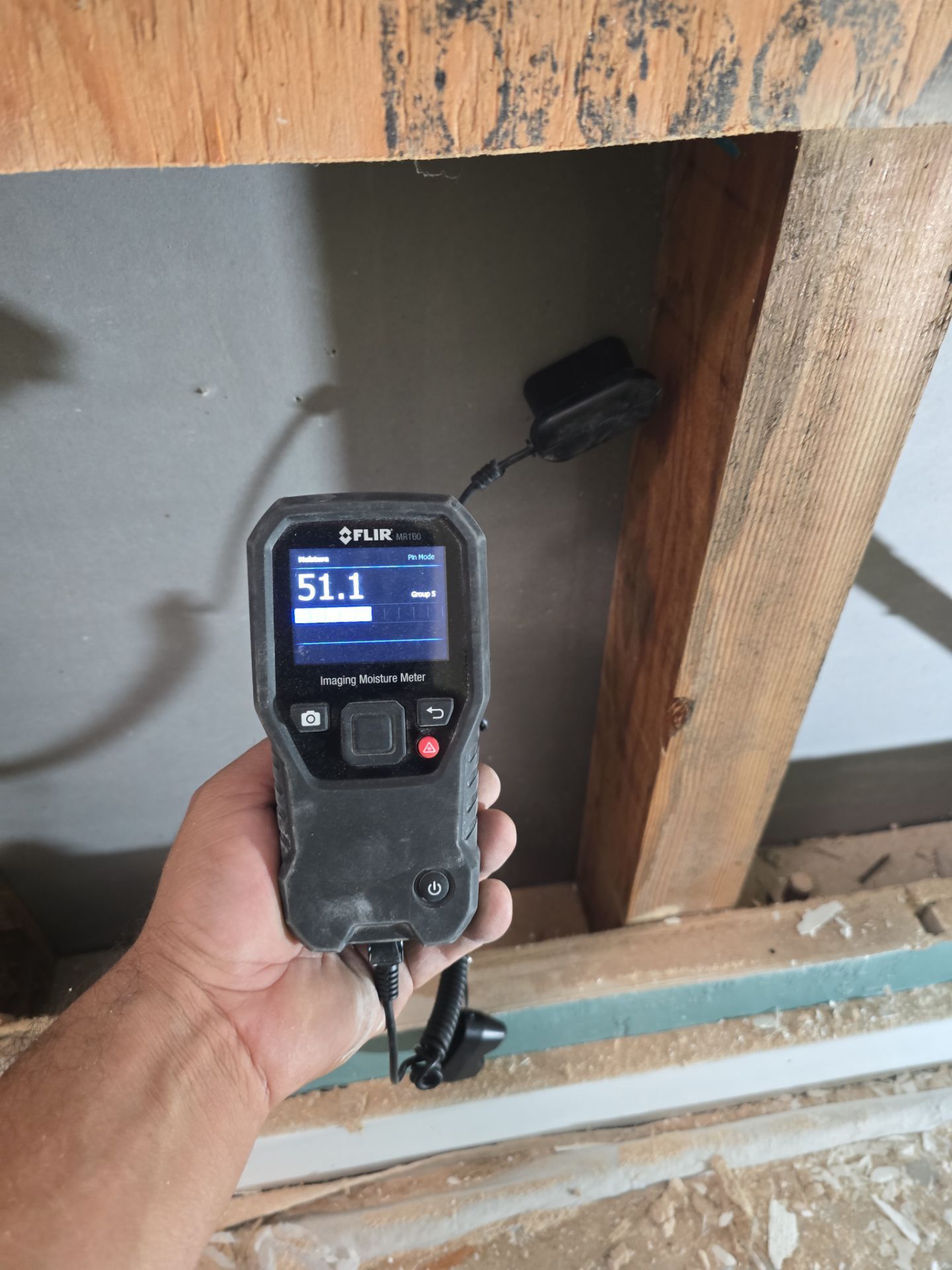 Handheld thermal camera aimed at a wall cavity with exposed wooden framing and insulation.