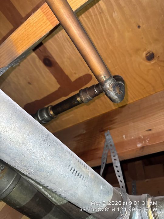 Copper plumbing pipes under wooden floor joists with a 90-degree elbow and metal hanger brace.