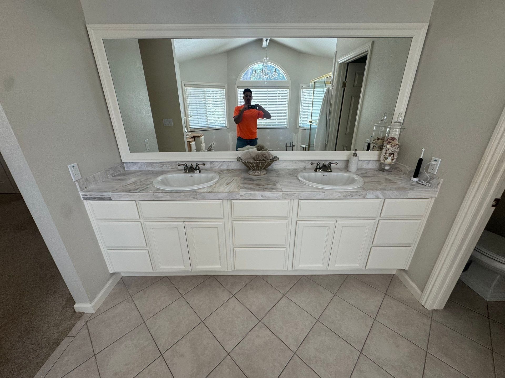 Bathroom vanity with double sinks, large mirror, and a person taking the photo reflected in it.