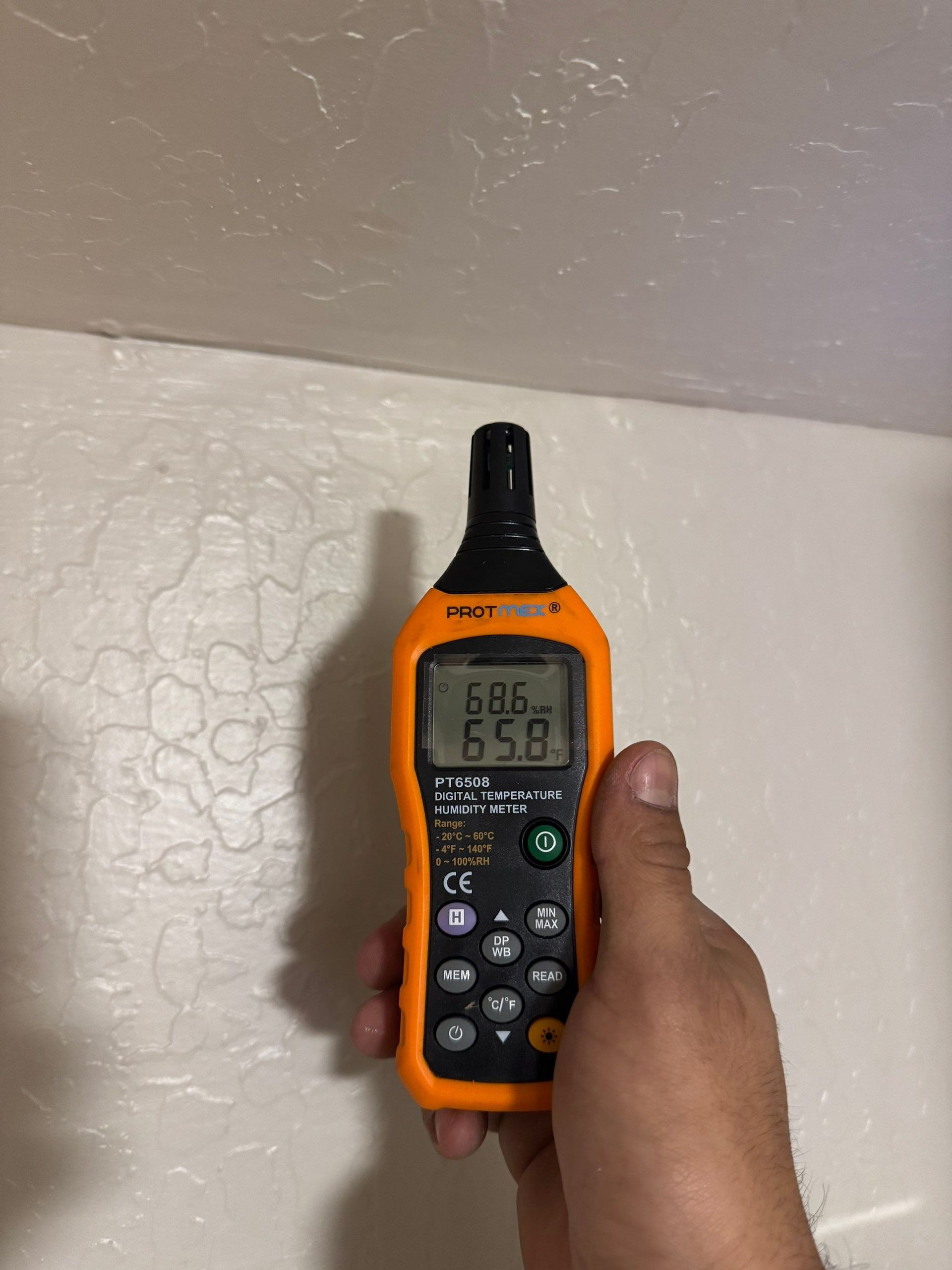 Hand holding an orange digital moisture meter near a white wall, showing readings on its display