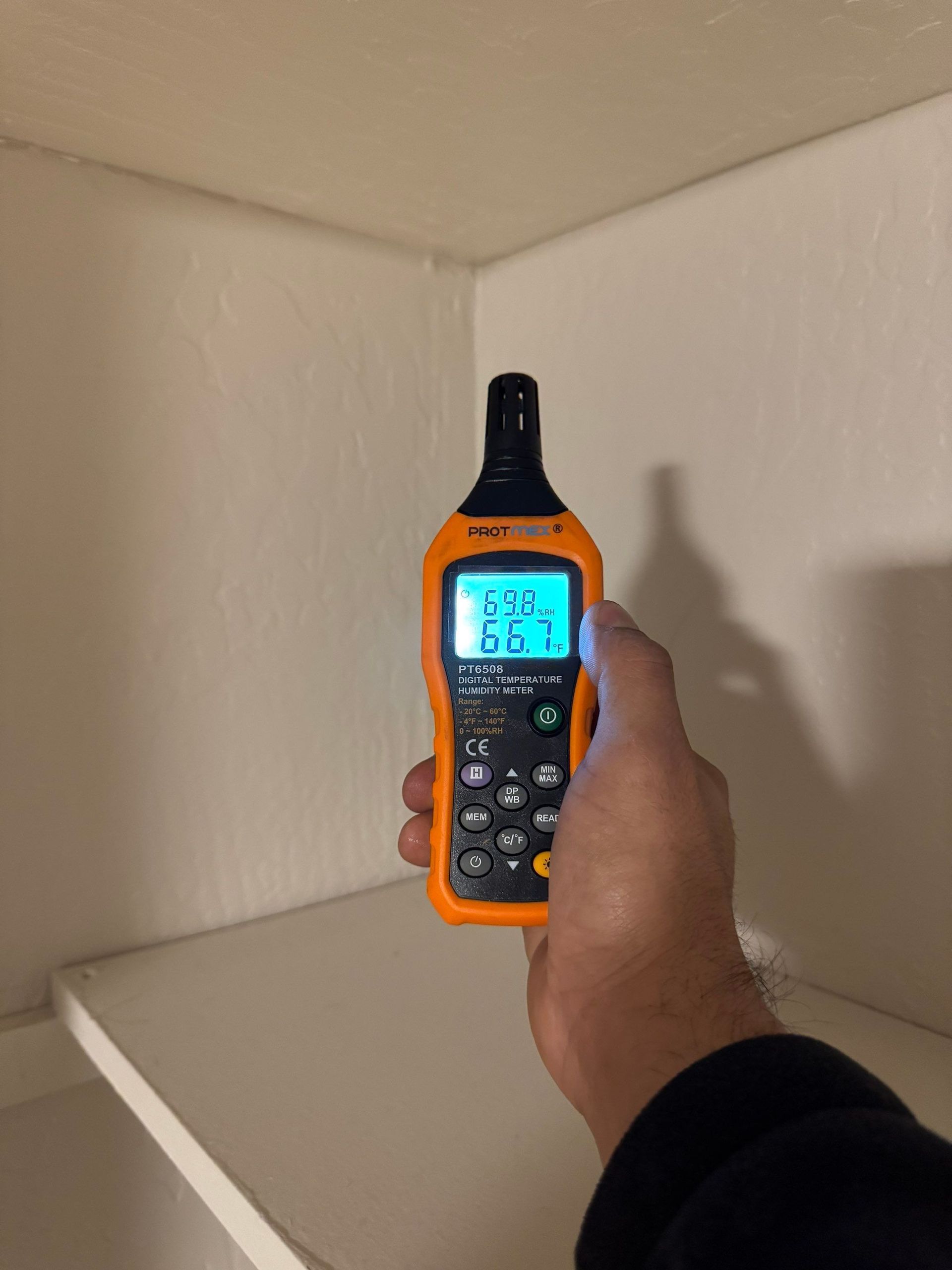 Hand holding a handheld meter toward a corner wall, with the screen lit blue.