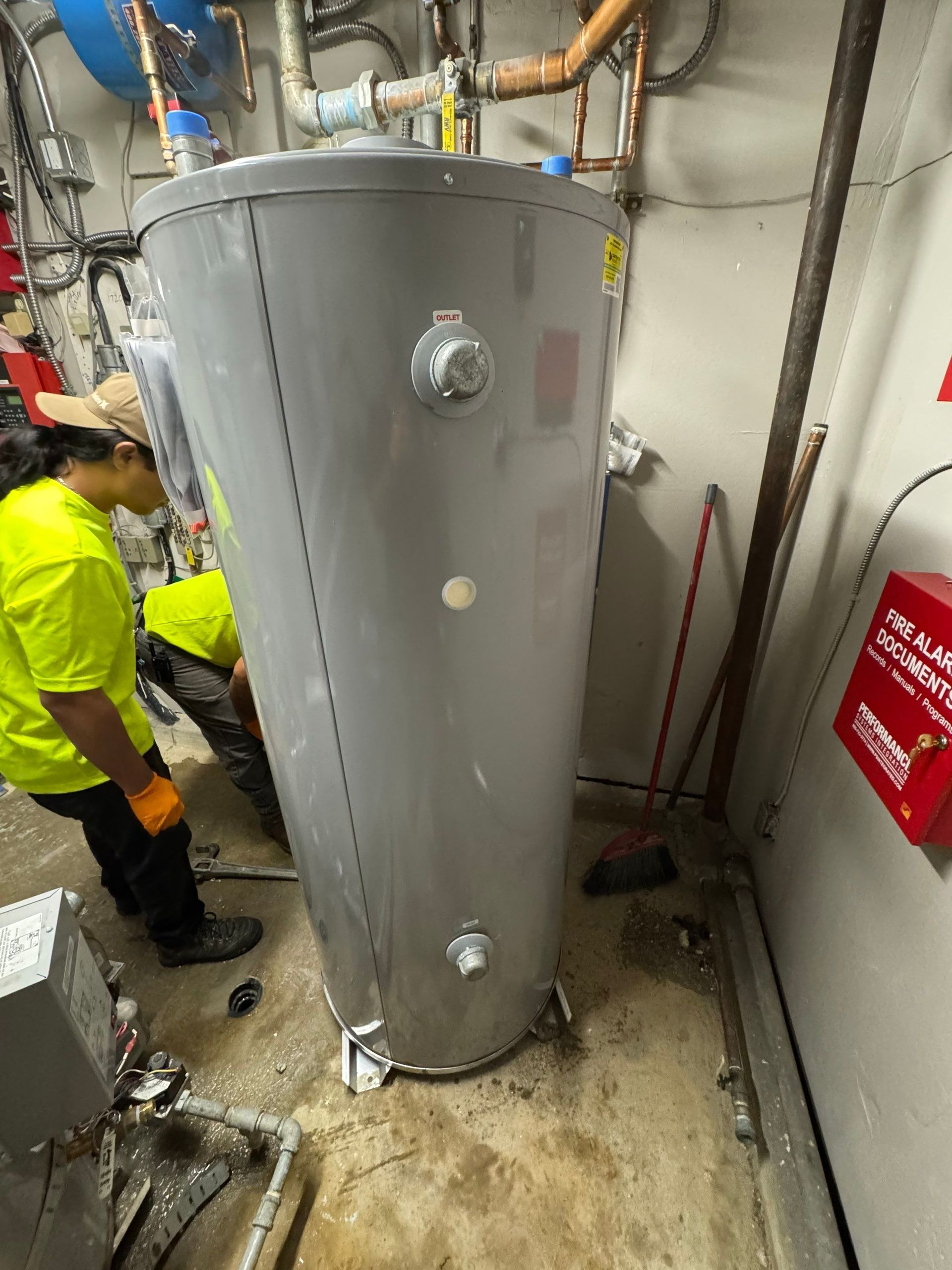 Gray industrial tank in a utility room, with a worker crouched beside it and pipes overhead.