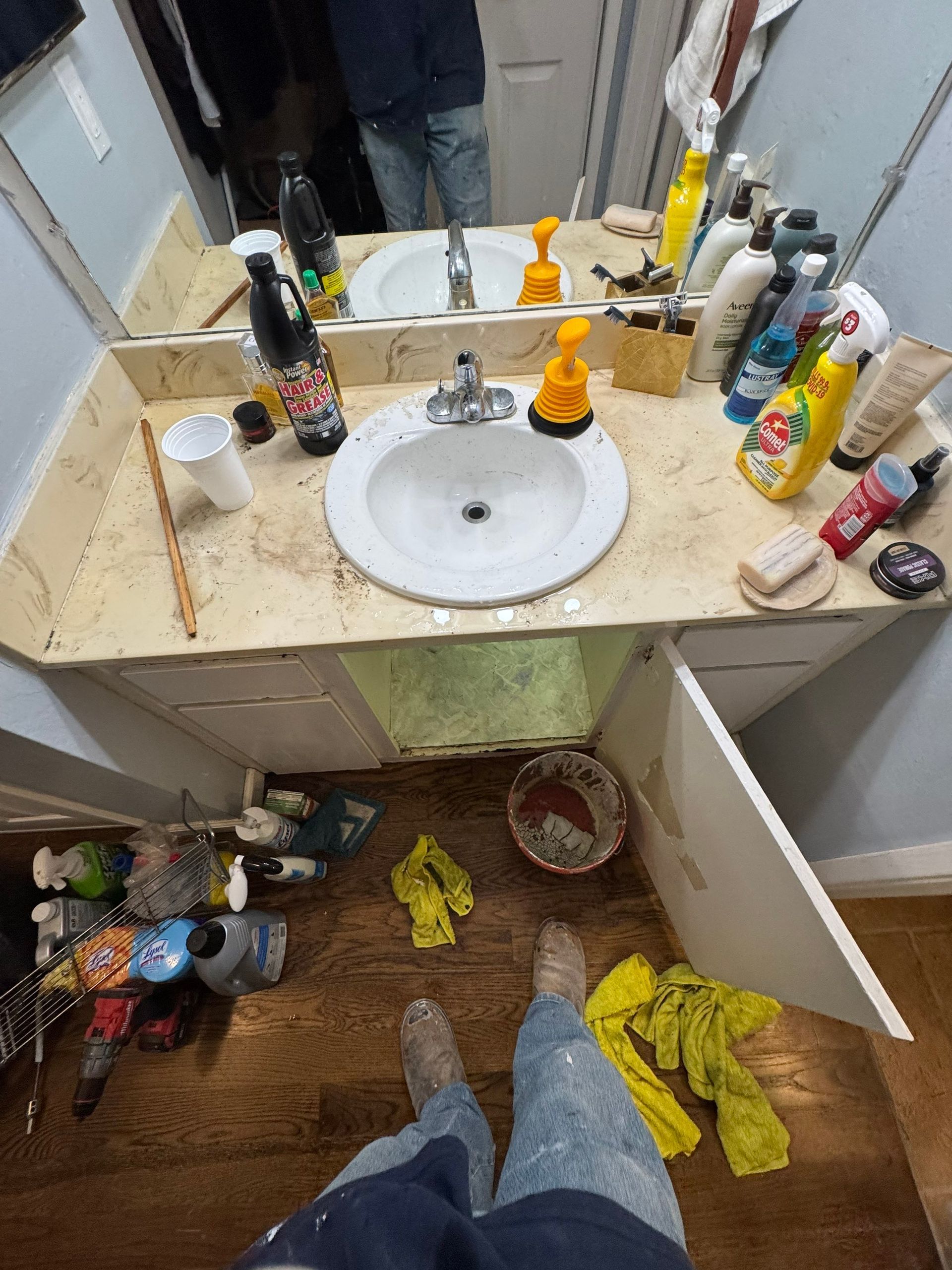 Messy bathroom vanity with a sink, scattered toiletries, and clutter on the floor.