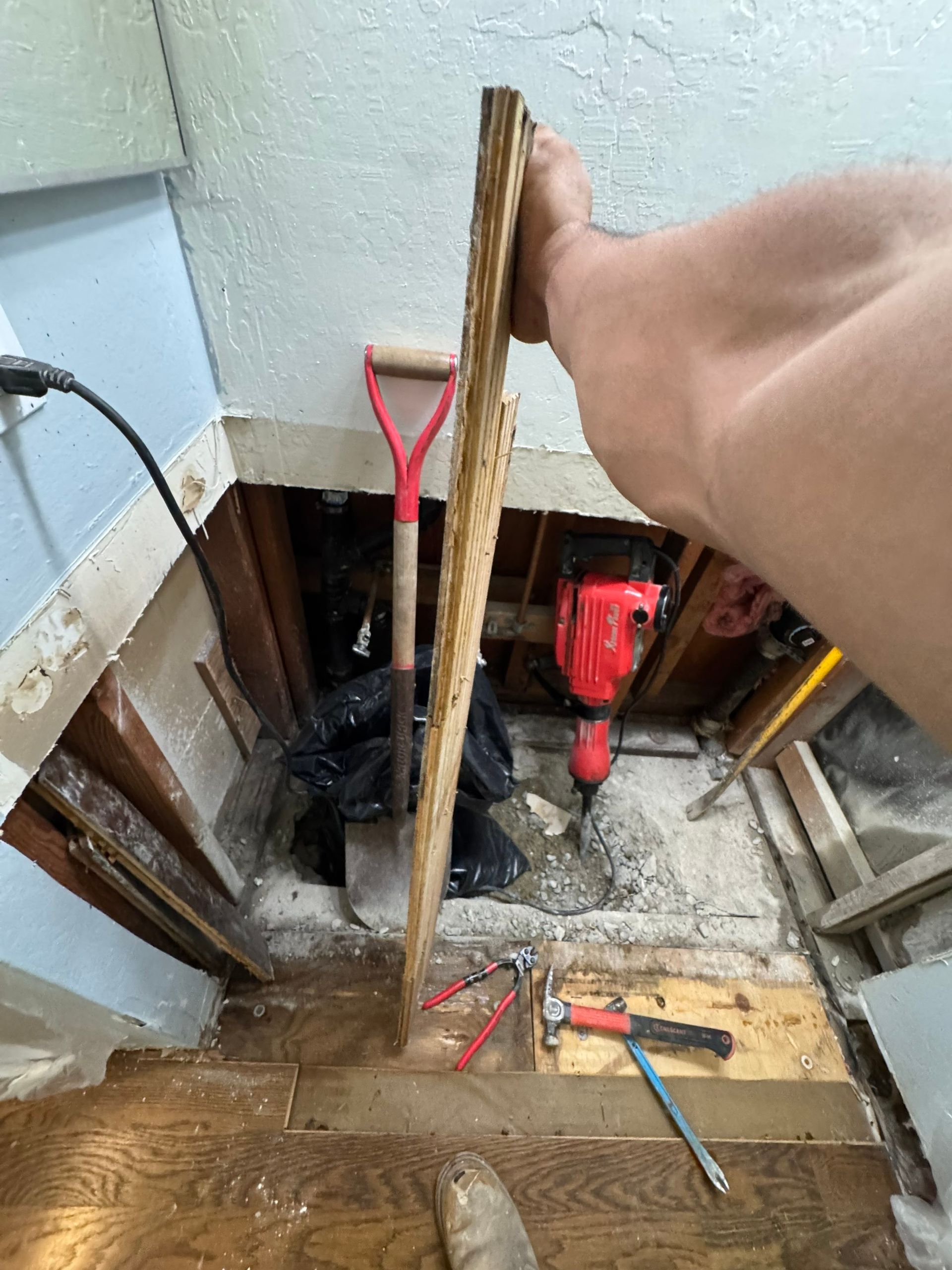 Worker leaning over a basement floor opening with tools and exposed pipes during home repair