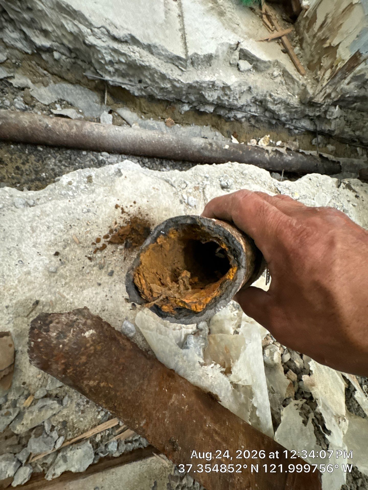 Hand holding a corroded metal pipe with orange rust inside amid rubble and broken concrete