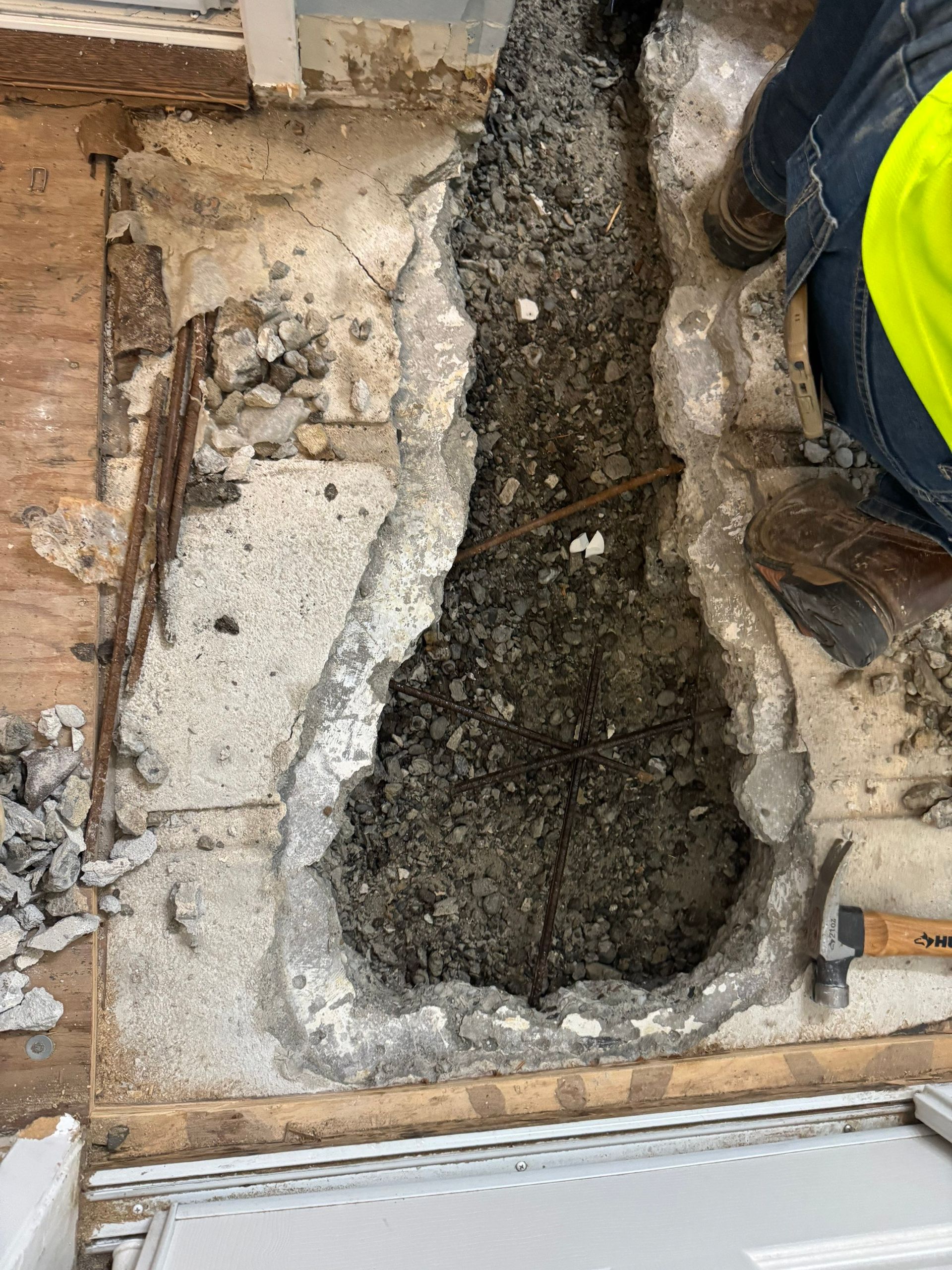 Excavated trench in a concrete floor beside exposed brick and rubble, with a worker’s boot at right.