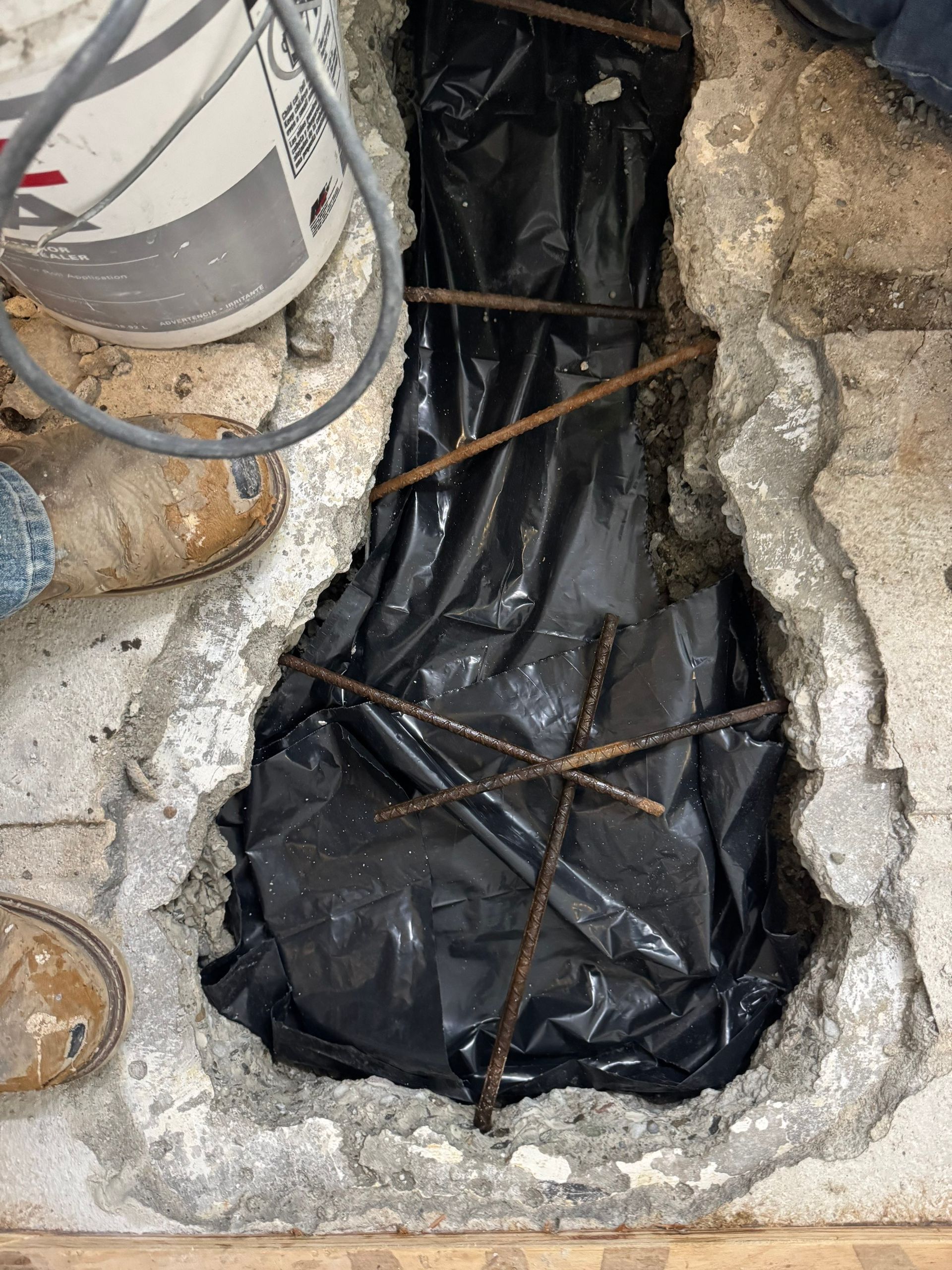 Broken concrete hole covered with black plastic and rebar, with boots visible on the left.