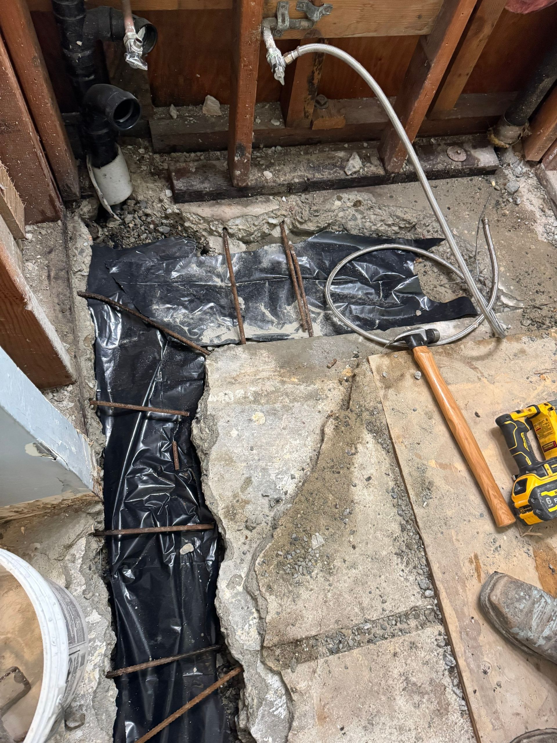 Exposed bathroom floor repair with black vapor barrier, pipes, and tools on concrete base