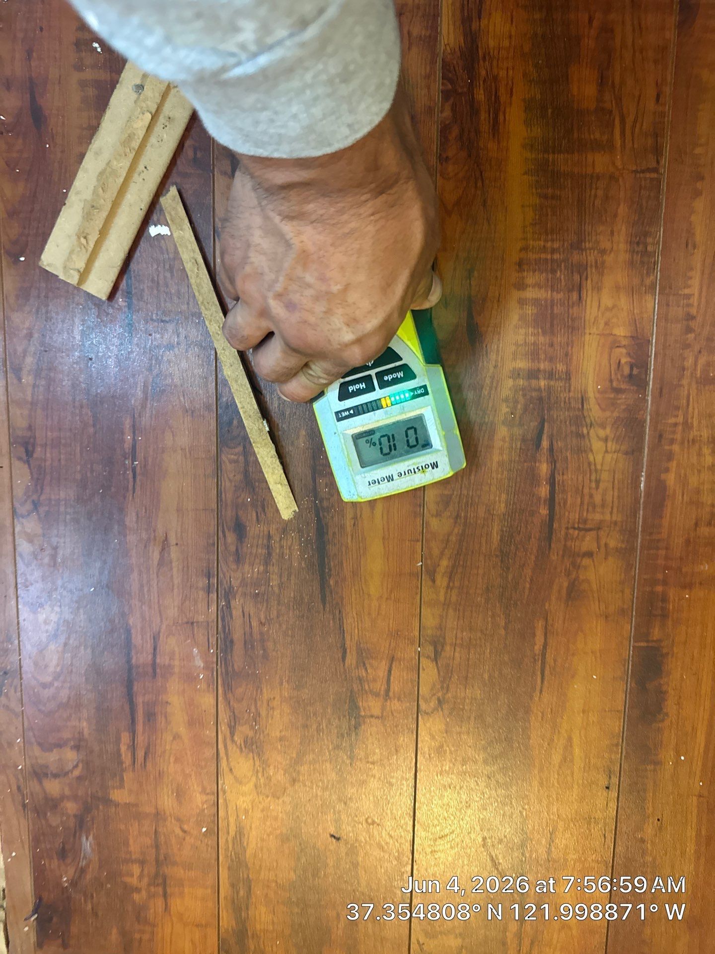 Hand holding a digital meter over a wooden floor with measuring sticks nearby