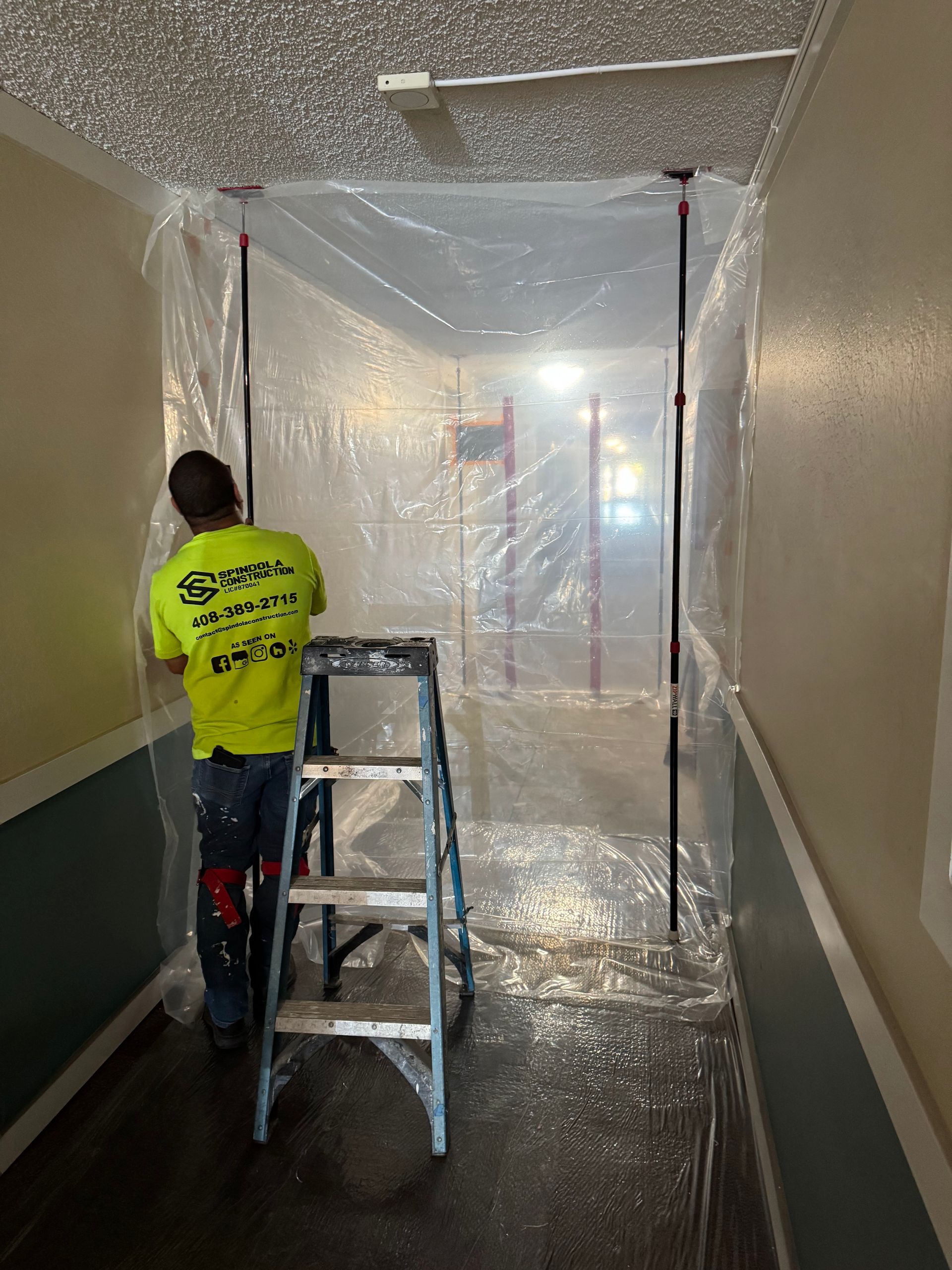 Worker on ladder sealing a hallway with clear plastic sheeting.
