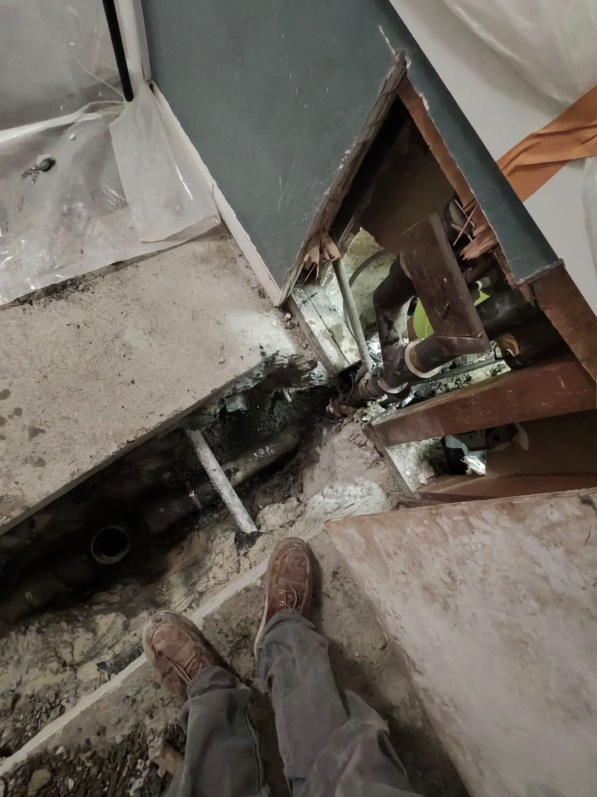 Damaged floor opening with exposed pipes and debris beside a person’s feet on a construction site