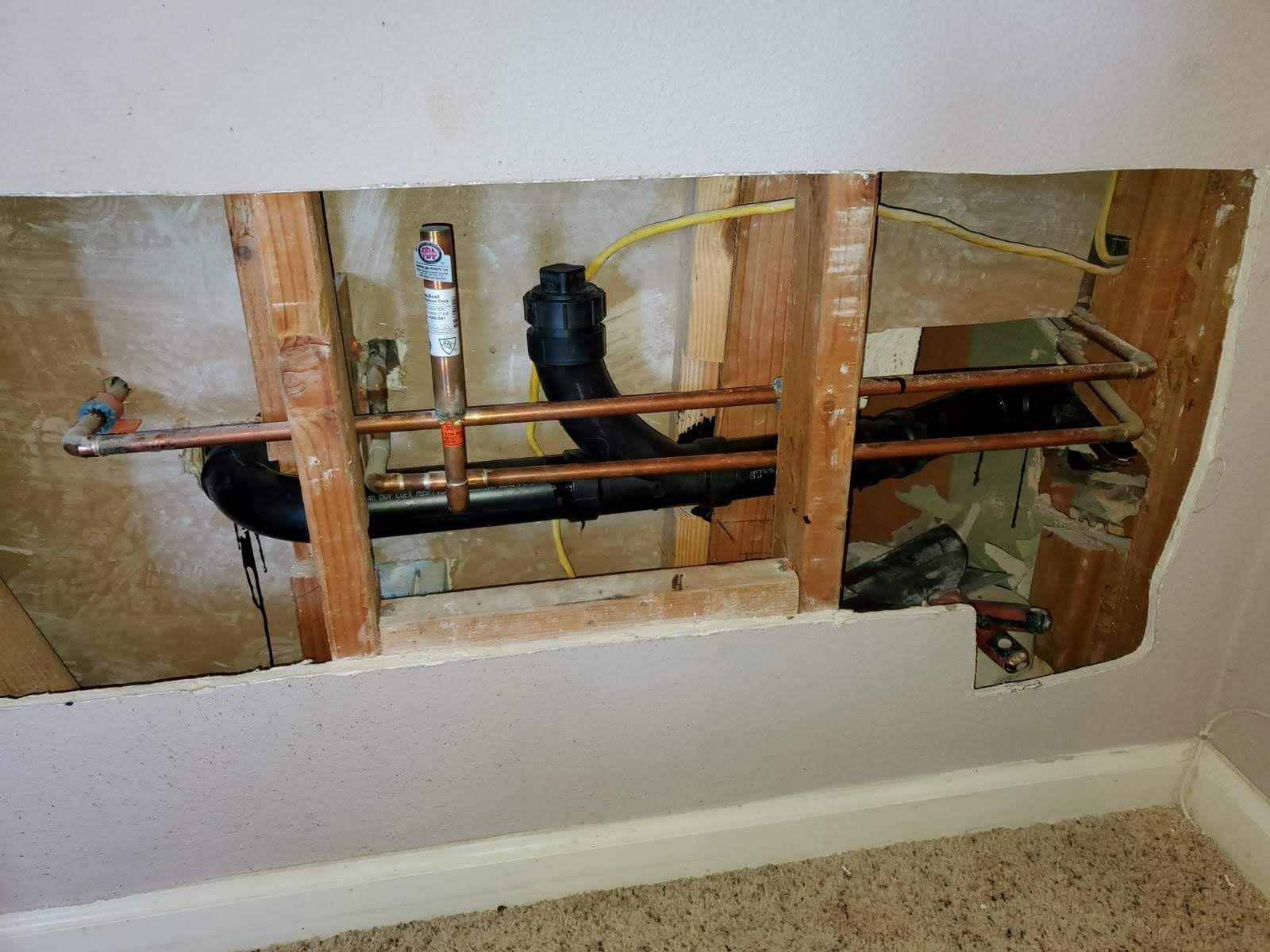 Exposed plumbing and pipes inside a wall opening above a baseboard, with wood framing and drywall cutout.