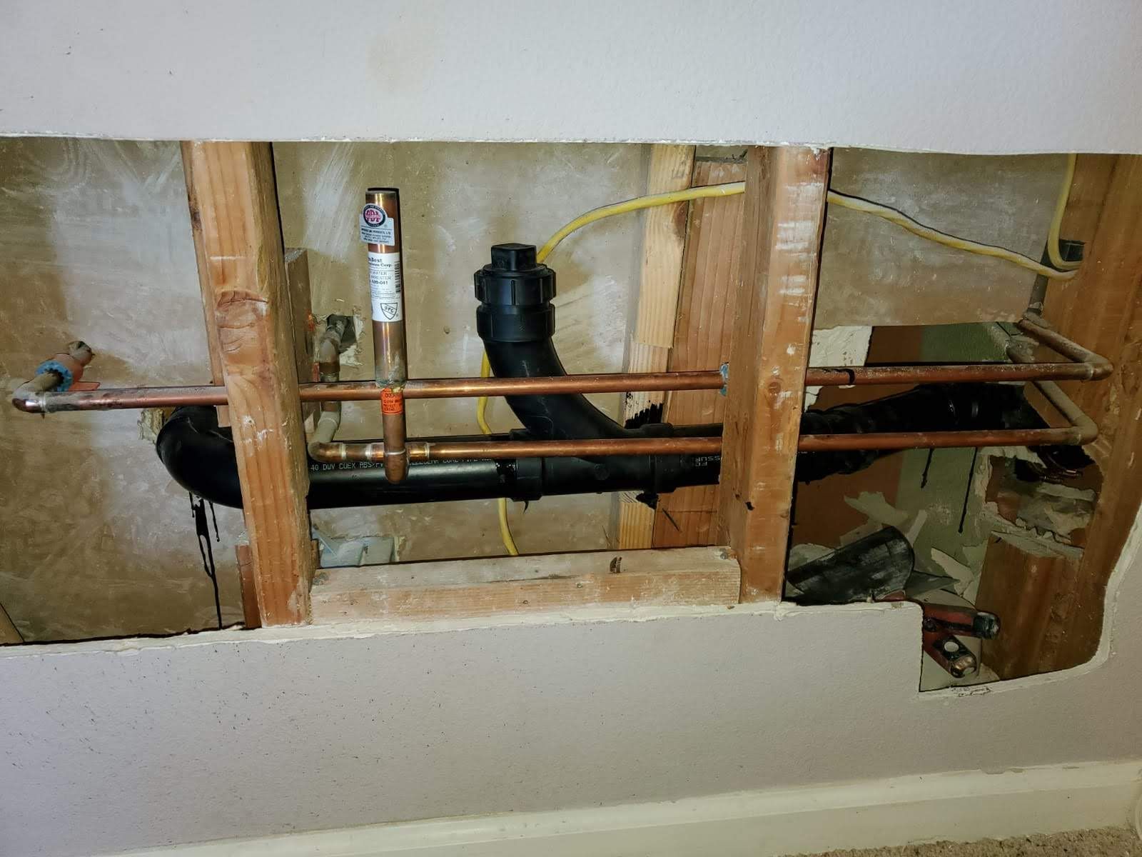 Open wall behind a toilet, showing plumbing pipes, wood studs, and exposed insulation.