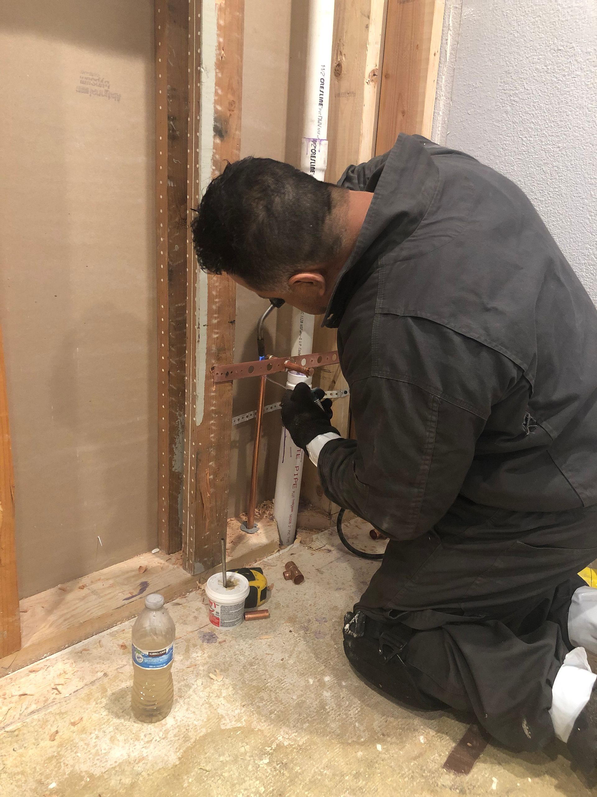 Worker kneeling beside exposed wall studs, using a tool on wiring during home renovation.
