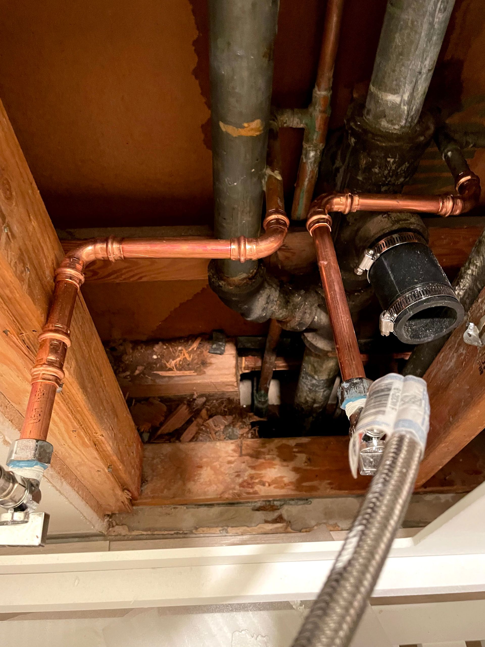 Exposed copper plumbing pipes and a braided hose under a sink or ceiling opening with water damage stains