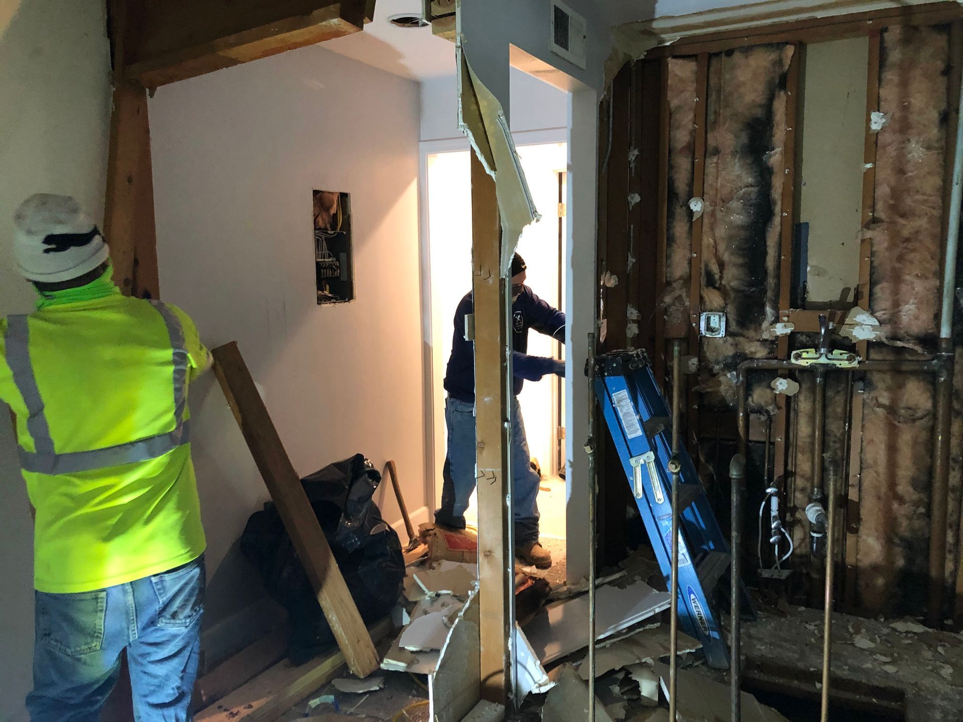 Worker in neon vest amid indoor construction debris, with exposed wall framing and tools.