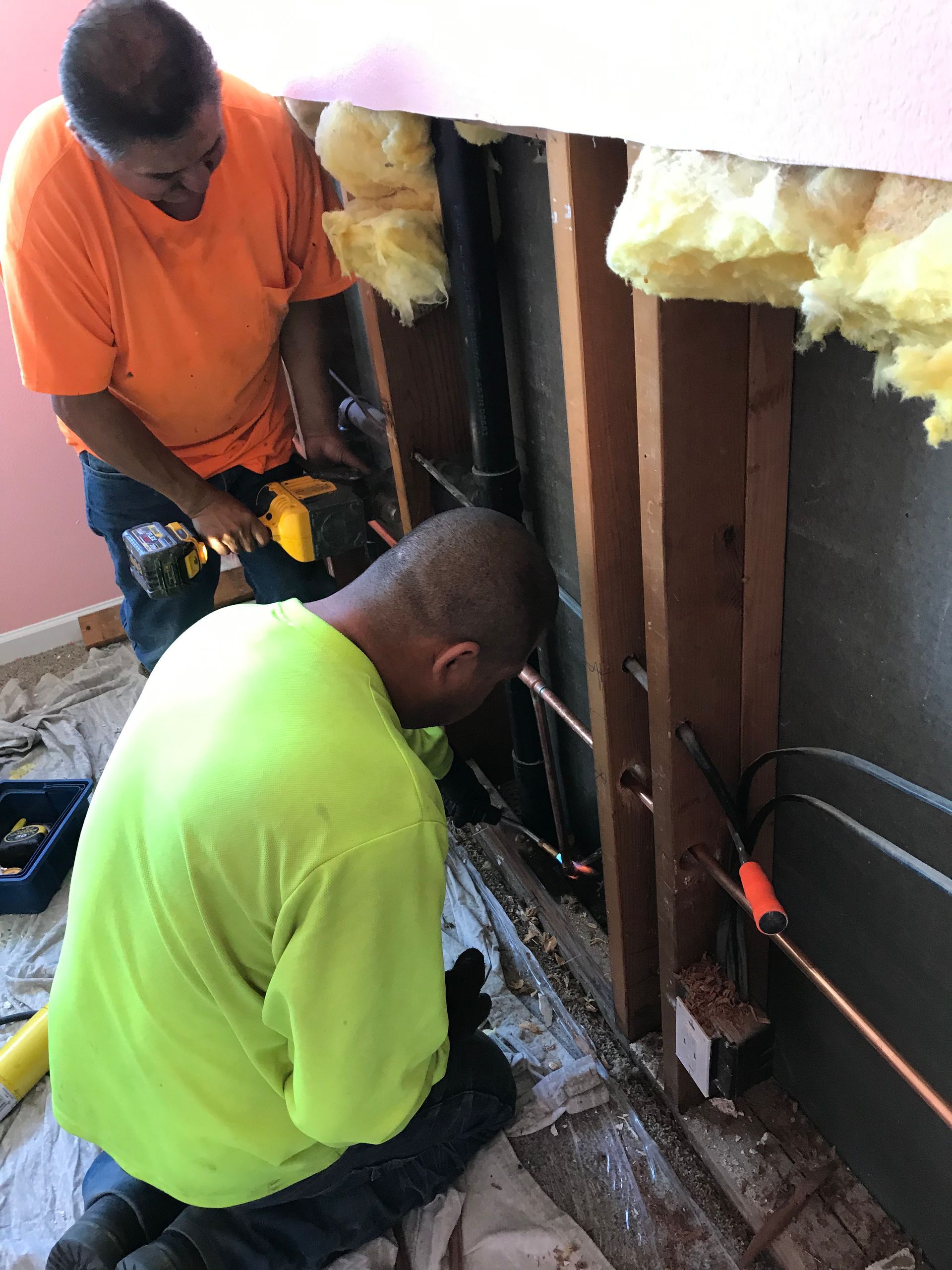 Two workers install wiring inside a wall cavity, one in orange and one in neon green.