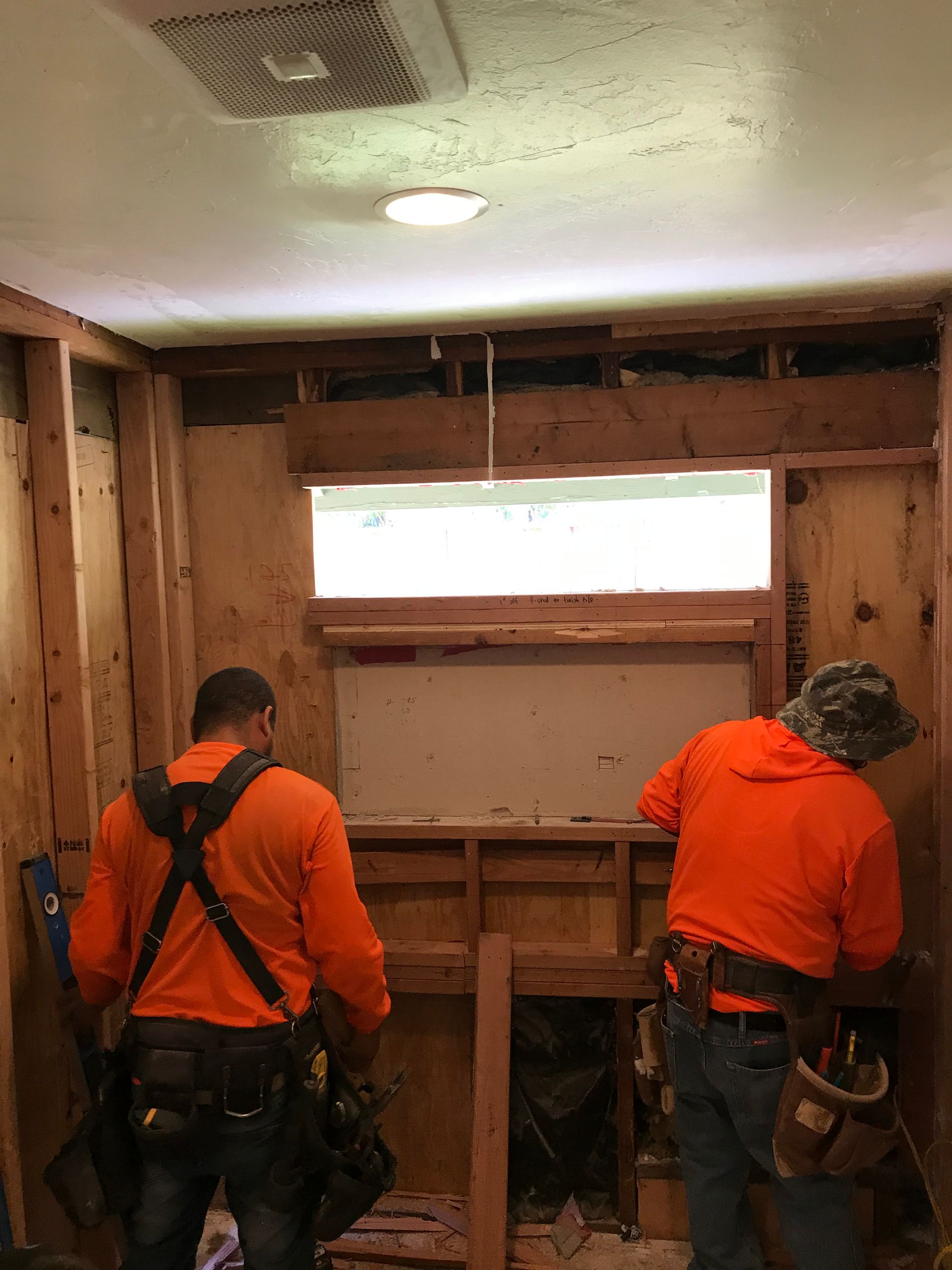 Two workers in orange shirts framing a wall inside a stripped room.