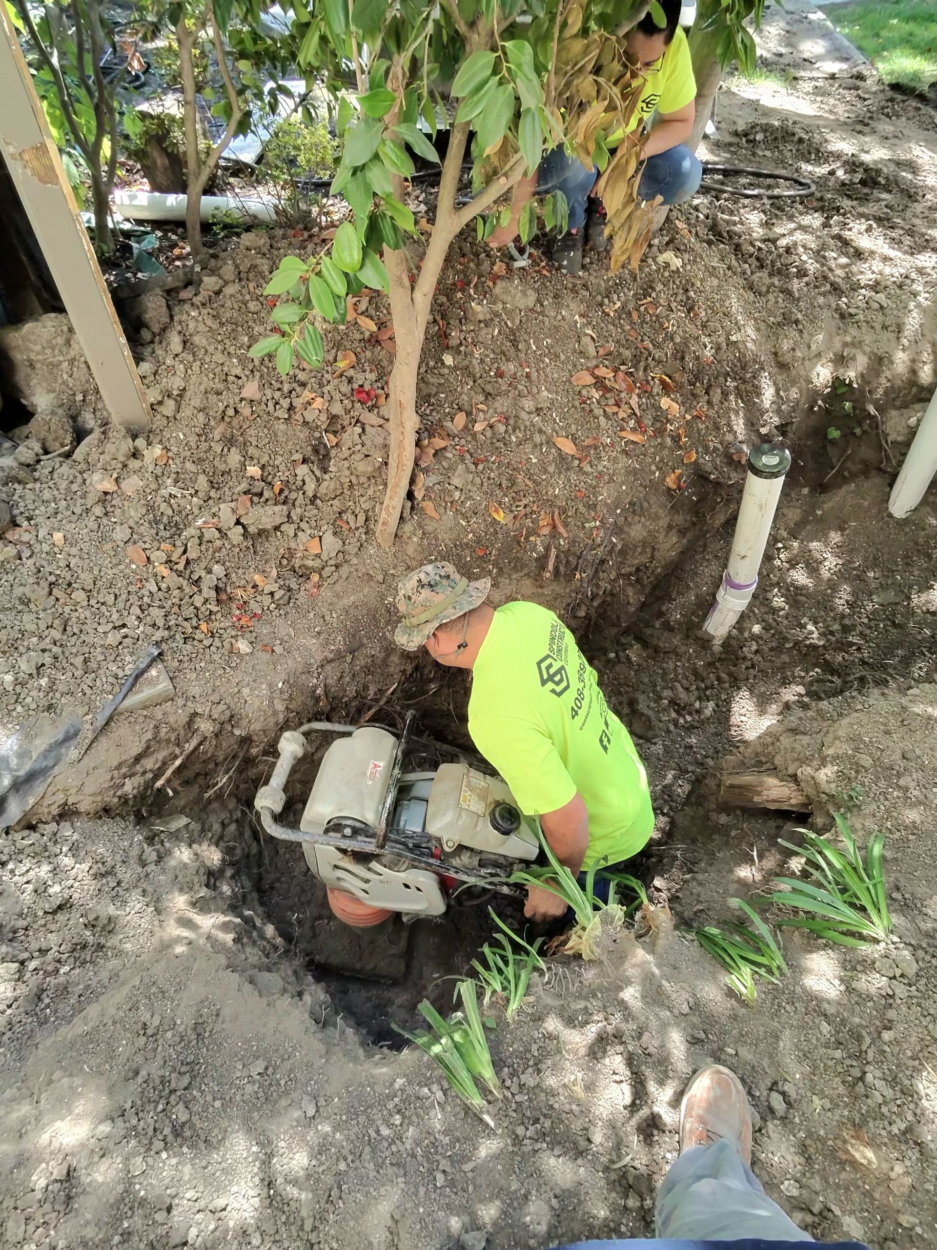 Person in neon green shirt working with a small machine in a dirt trench under trees