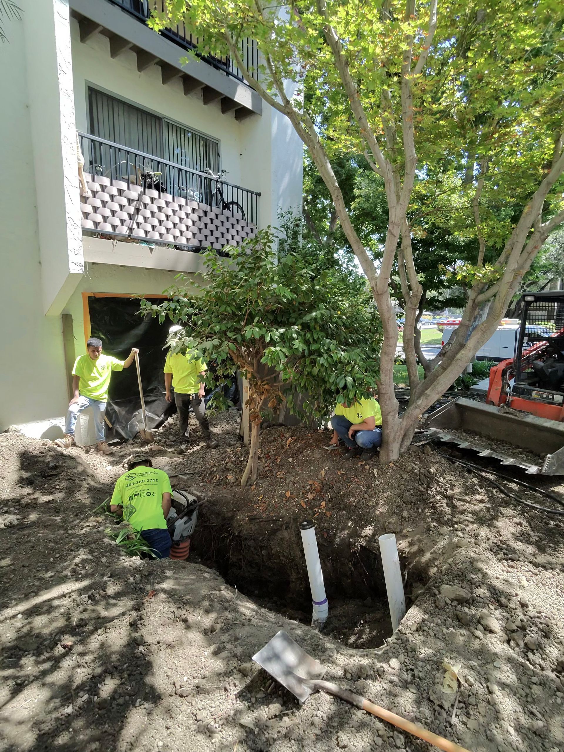 Workers digging a muddy excavation beside a building and trees, with a balcony overhead