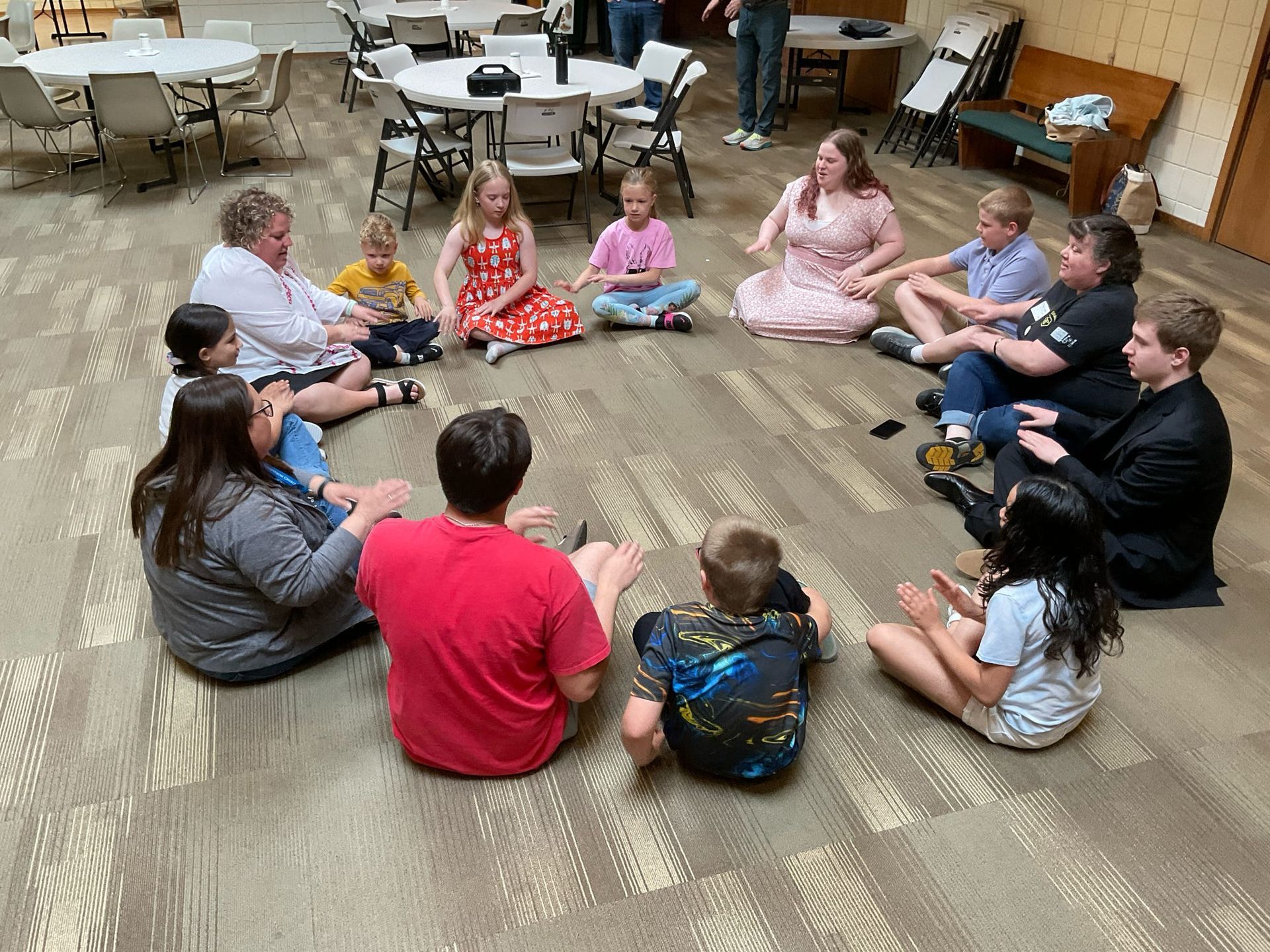 A group of children and teens are sitting in a circle on the floor for singing prayer.