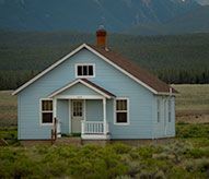A small blue house with a porch in a field with mountains in the background.