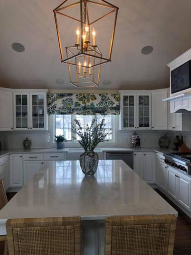 Bright white kitchen with a large island, wicker stools, glass cabinets, and geometric pendant lights.
