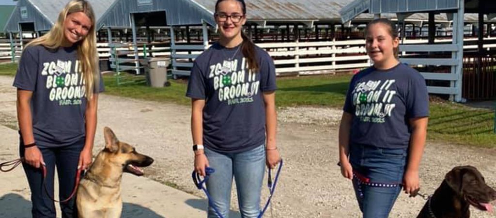 Three people wearing matching navy shirts stand outside with two dogs on leashes, one a German Shepherd, one a brown dog.