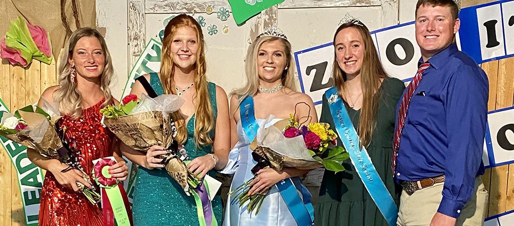 Four people stand in a line holding flower bouquets, with a man standing on the far right in front of a celebratory backdrop.