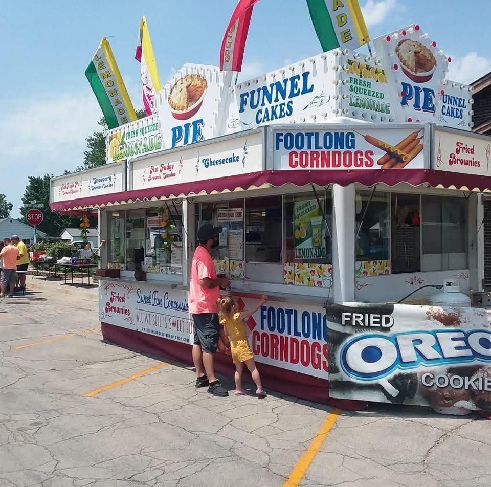 A person and a child stand at a colorful carnival food stand selling funnel cakes, corn dogs, and fried Oreos.