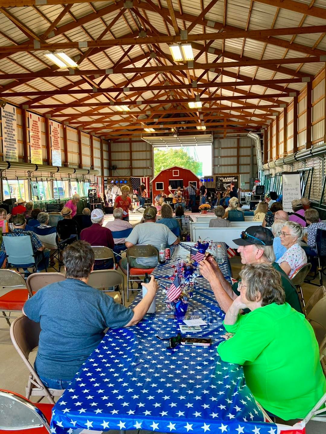 People sit at tables covered in star-patterned tablecloths inside a rustic, open-air wooden barn at a social event.