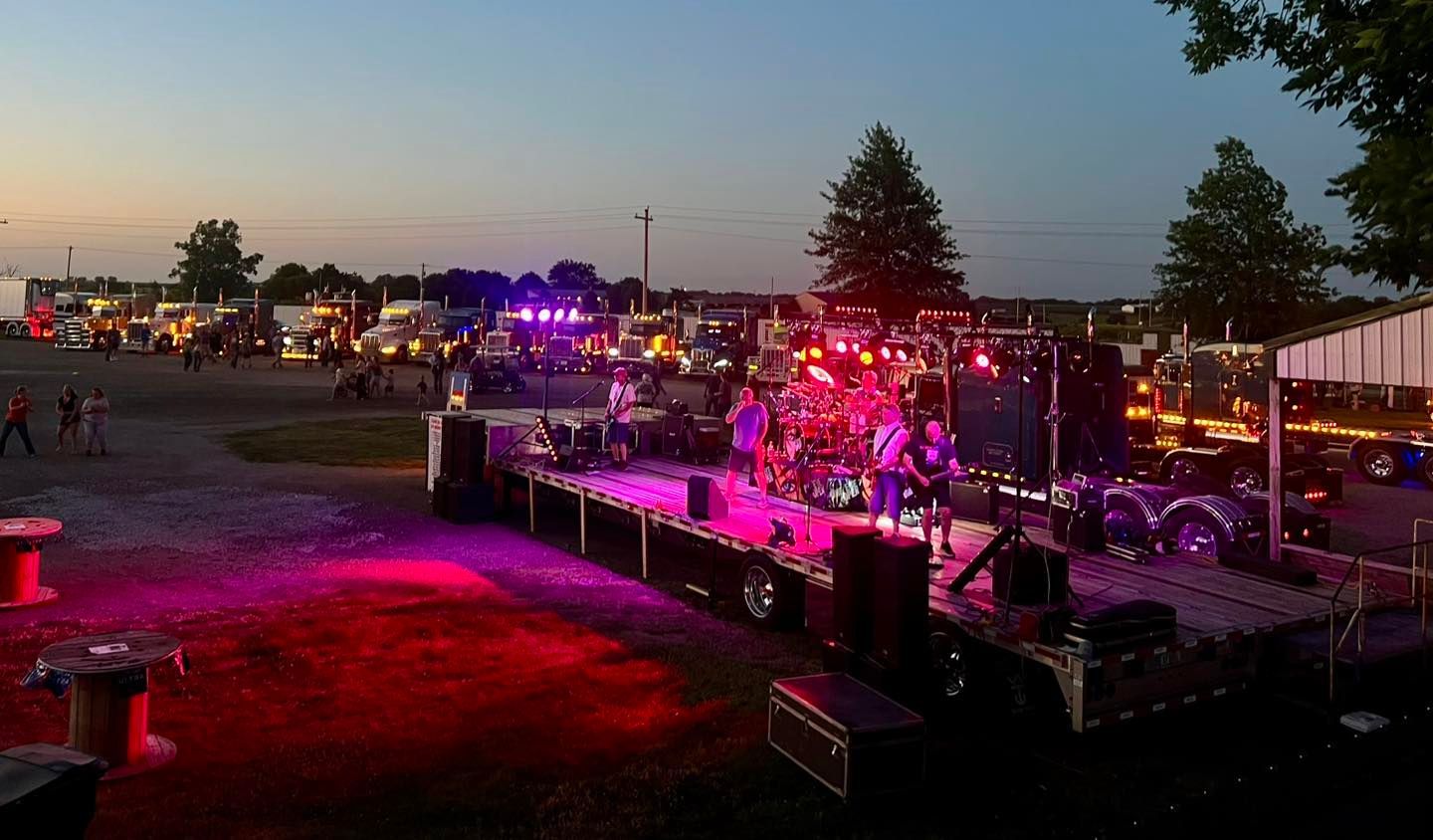 A band performs on a trailer-stage illuminated by purple lights at an outdoor event filled with semi-trucks at dusk.