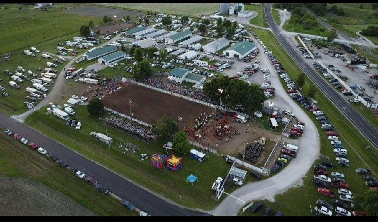 Aerial view of a rodeo arena surrounded by parked vehicles, crowds of people, and buildings in a rural setting.