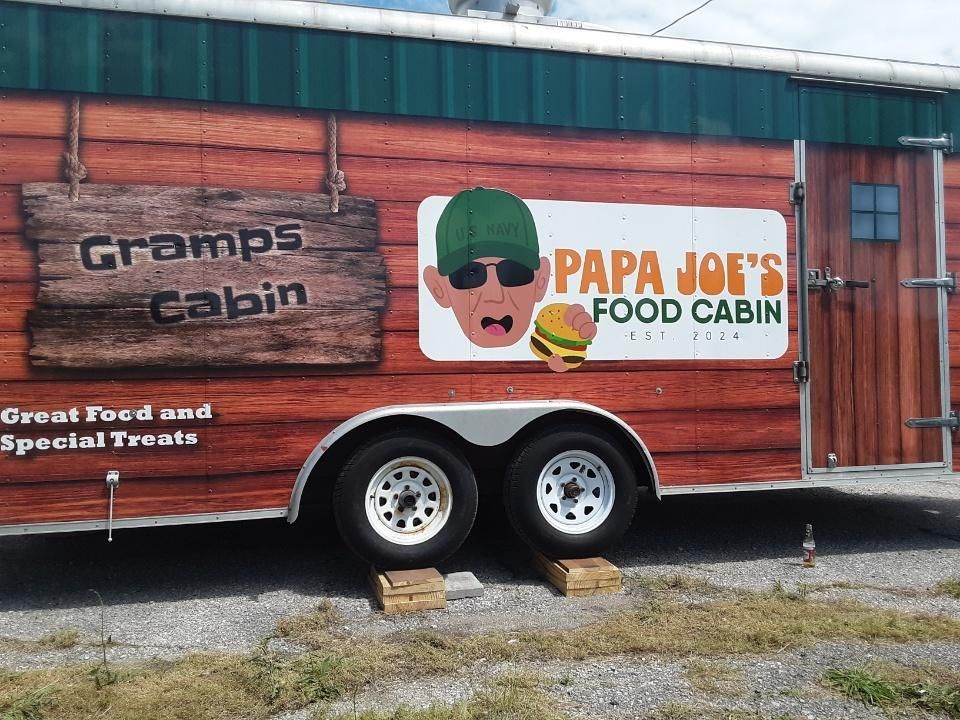 Side view of a 'Papa Joe's Food Cabin' food trailer with wood-patterned siding, a graphic of a man, and a burger icon.