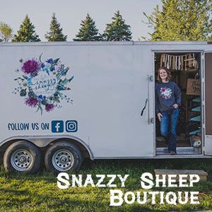 A woman stands in the open doorway of a white mobile boutique trailer labeled 'Snazzy Sheep Boutique.'