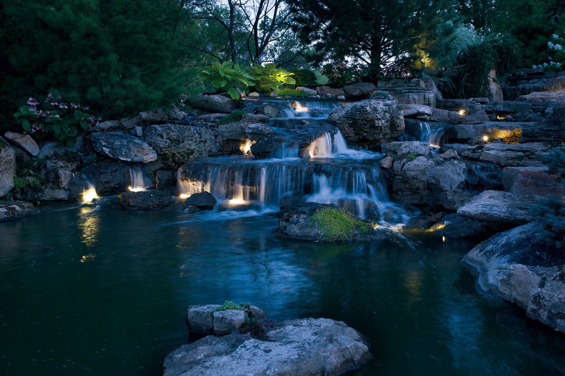 Large Ohio water feature with tier cascading waterfalls and large rocks at night with lights