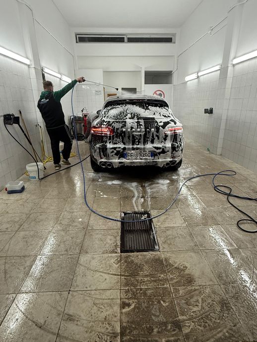 Una persona lava un'auto nera con la schiuma di sapone; una mano con un guanto rosa pulisce il cofano e il faro.
