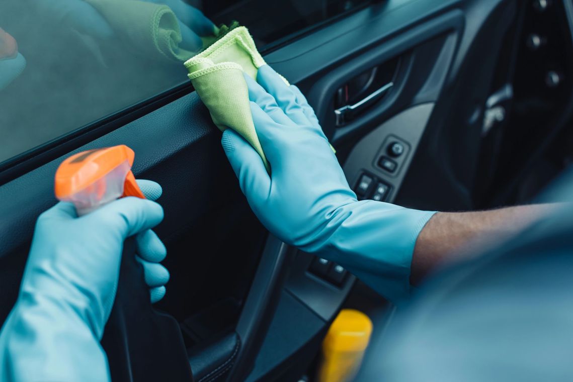 Mani guantate che puliscono l'interno di un'auto con un flacone spray e dei panni.